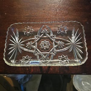 May include: A clear, rectangular glass serving tray with a decorative starburst and star pattern. The tray has a scalloped edge and is designed for serving appetizers or desserts. The tray is on a dark brown wooden surface.
