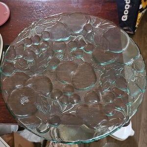 May include: A clear glass serving platter with a fruit design. The round platter features embossed grapes, pears, and leaves. The platter is approximately 12 inches in diameter. The glass has a slight green tint.