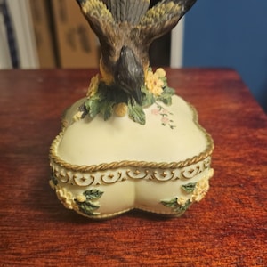 May include: A white ceramic mug with a black handle and the text "But First, Coffee" in a bold, black font. The mug is set against a blurred background, suggesting a cozy setting.