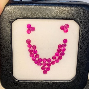 Pode incluir: Uma caixa preta exibe uma disposição de pequenas pedras preciosas redondas rosa brilhante em forma de colar. As pedras são dispostas sobre um fundo branco, criando um contraste marcante. A disposição assemelha-se a um rosto sorridente.