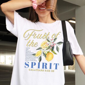 Op de afbeelding: Wit T-shirt met de tekst "Fruit of the Spirit" in geel en blauw, met een citroen- en bloemenontwerp. Het shirt vermeldt ook deugden als liefde, vreugde, vrede en geduld. De tekst "Galaten 5:22-23" is ook opgenomen.