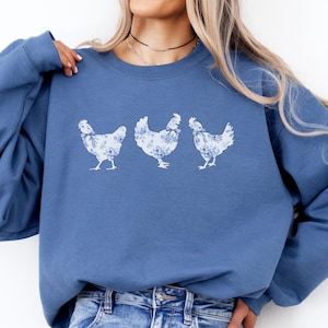 Może przedstawiać: Niebieska bluza w kolorze indygo z trzema białymi i niebieskimi kwiatowymi ilustracjami kur. Bluza z okrągłym dekoltem wykonana jest z miękkiego materiału i ma wygodny krój. Tekst "indigo blue" znajduje się po lewej stronie u dołu.