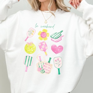 Könnte beinhalten: Weißes Sweatshirt mit einem farbenfrohen Grafikdesign mit Pickleball-Schlägern, Blumen und Herzen. Der Satz "le weekend" ist kursiv geschrieben. Das Wort "white" befindet sich in der unteren rechten Ecke.