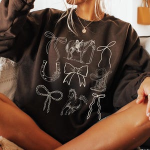 May include: Dark chocolate-colored sweatshirt featuring white line drawings of cowboy-themed elements. The design includes a horse and rider, bows, a horseshoe, a cowboy boot, and a cowboy hat. The text "dark chocolate" is visible at the bottom right.