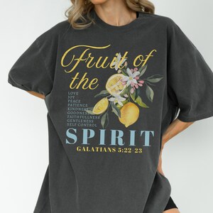 Op de afbeelding: Donkergrijs T-shirt met de tekst "Fruit of the Spirit" in gele letters, met een citroen- en bloemenontwerp. Het shirt vermeldt deugden als liefde, vreugde en vrede, met de Bijbelverwijzing Galaten 5:22-23.