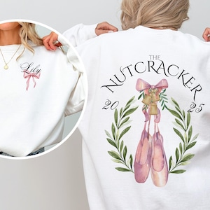 Könnte beinhalten: Weißes Sweatshirt mit einem Ballettthema. Auf der Rückseite befindet sich eine Aquarellillustration von Spitzenschuhen, Glocken und einer Schleife mit dem Text "The Nutcracker 2025". Auf der Vorderseite steht der Name "Lily" und eine rosa Schleife.