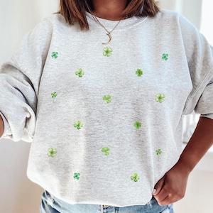 May include: Light grey sweatshirt decorated with small green shamrock motifs. This long-sleeved top has a crew neck and a loose fit. The text "ash gray" is visible at the bottom left. A comfortable garment for everyday wear.