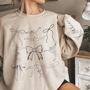 May include: Beige sweatshirt featuring black line art of gymnasts in various poses and bow illustrations. The sleeve has the word "love" in cursive with bow details. A comfortable, casual top for those who love gymnastics.