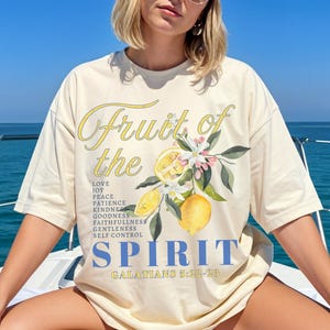 Op de afbeelding: Ivoor kleurig T-shirt met de tekst "Fruit of the Spirit" in gele letters en blauwe blokletters. Het shirt heeft een citroen en bloemen grafische afbeelding. De tekst bevat een lijst met deugden en de Bijbeltekst Galaten 5:22-23.