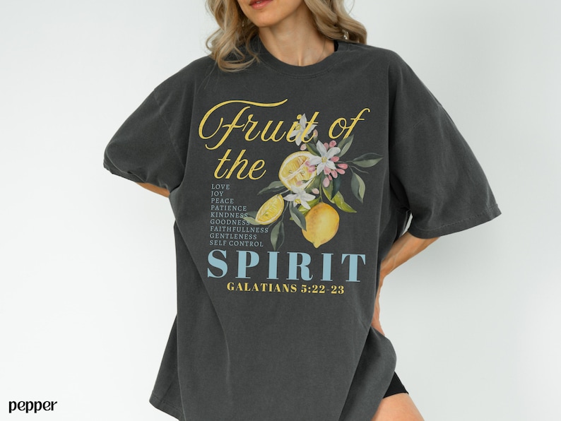 Op de afbeelding: Donkergrijs T-shirt met de tekst "Fruit of the Spirit" in gele letters, met een citroen- en bloemenontwerp. Het shirt vermeldt deugden als liefde, vreugde en vrede, met de Bijbelverwijzing Galaten 5:22-23.