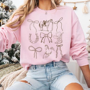 May include: Light pink sweatshirt with brown line drawings of cowboy-themed elements. The design includes a horse, cowboy hat, horseshoe, and bows. The words "light pink" are at the bottom left of the image.