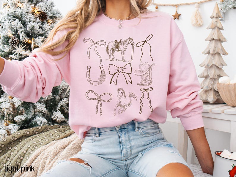 May include: Light pink sweatshirt with brown line drawings of cowboy-themed elements. The design includes a horse, cowboy hat, horseshoe, and bows. The words "light pink" are at the bottom left of the image.