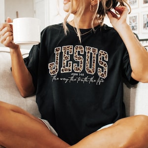 May include: Black t-shirt with the word "JESUS" in a leopard print design. The text "John 14:6" and "the way the truth the life" are below the word "JESUS". A white mug is held in the left hand.