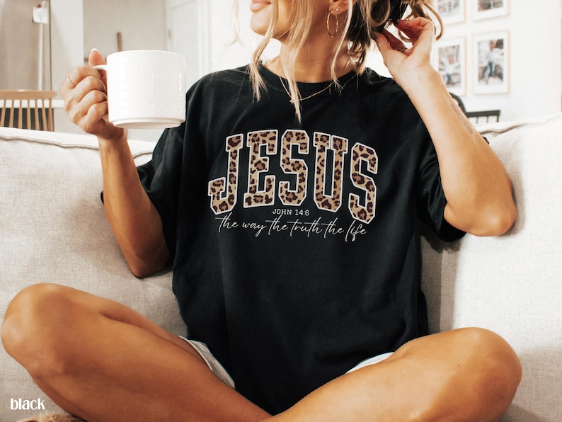 May include: Black t-shirt with the word "JESUS" in a leopard print design. The text "John 14:6" and "the way the truth the life" are below the word "JESUS". A white mug is held in the left hand.