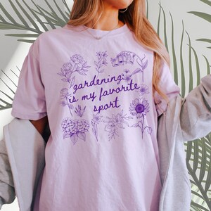 May include: A light purple t-shirt with the text "gardening is my favorite sport" in a purple floral design. The shirt is a soft, comfortable material.