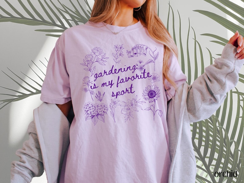 May include: A light purple t-shirt with the text "gardening is my favorite sport" in a purple floral design. The shirt is a soft, comfortable material.