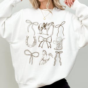 May include: White sweatshirt featuring a collection of brown line art illustrations. The design includes a horse with a saddle, a cowboy boot, a horseshoe, bows, and a cowboy hat. The word "white" is printed in the bottom right corner.