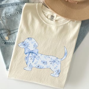 May include: Ivory-colored t-shirt featuring a blue floral dachshund design with a bow. The shirt is laid on top of a pair of light blue jeans and a tan hat. A silver necklace with a pendant is visible.