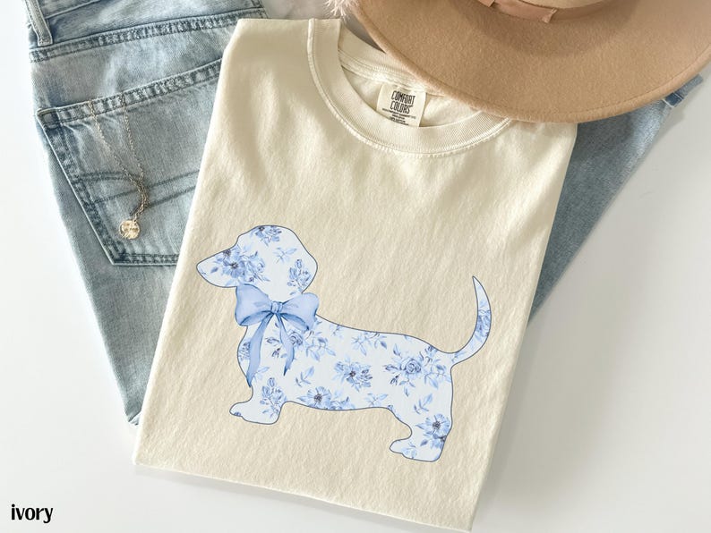 May include: Ivory-colored t-shirt featuring a blue floral dachshund design with a bow. The shirt is laid on top of a pair of light blue jeans and a tan hat. A silver necklace with a pendant is visible.