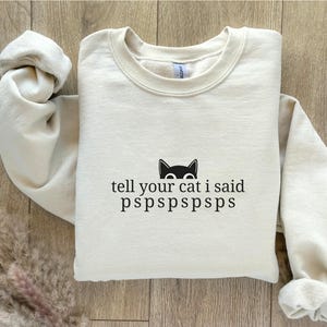 Puede incluir: Sudadera de color arena con el texto "tell your cat i said pspspspsps" y un gráfico de gato negro. La sudadera está hecha de un material suave y tiene cuello redondo.