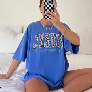 May include: A blue t-shirt with the word "JESUS" in a leopard print design across the chest. The text "the way the truth the life" is below the word "JESUS". The shirt is a relaxed fit.