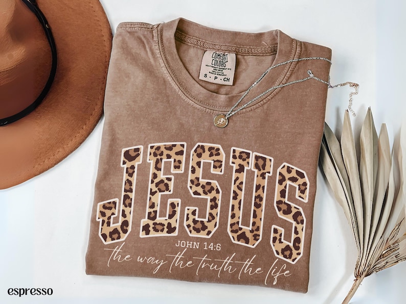 May include: Espresso-colored t-shirt with the word "JESUS" in a leopard print design. The text "JOHN 14:6 the way the truth the life" is printed below the word. A silver necklace and a brown hat are also in the image.