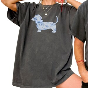 May include: Dark gray t-shirt featuring a light blue dachshund illustration with a floral pattern and a bow. The word "pepper" is printed in the lower left corner of the image.
