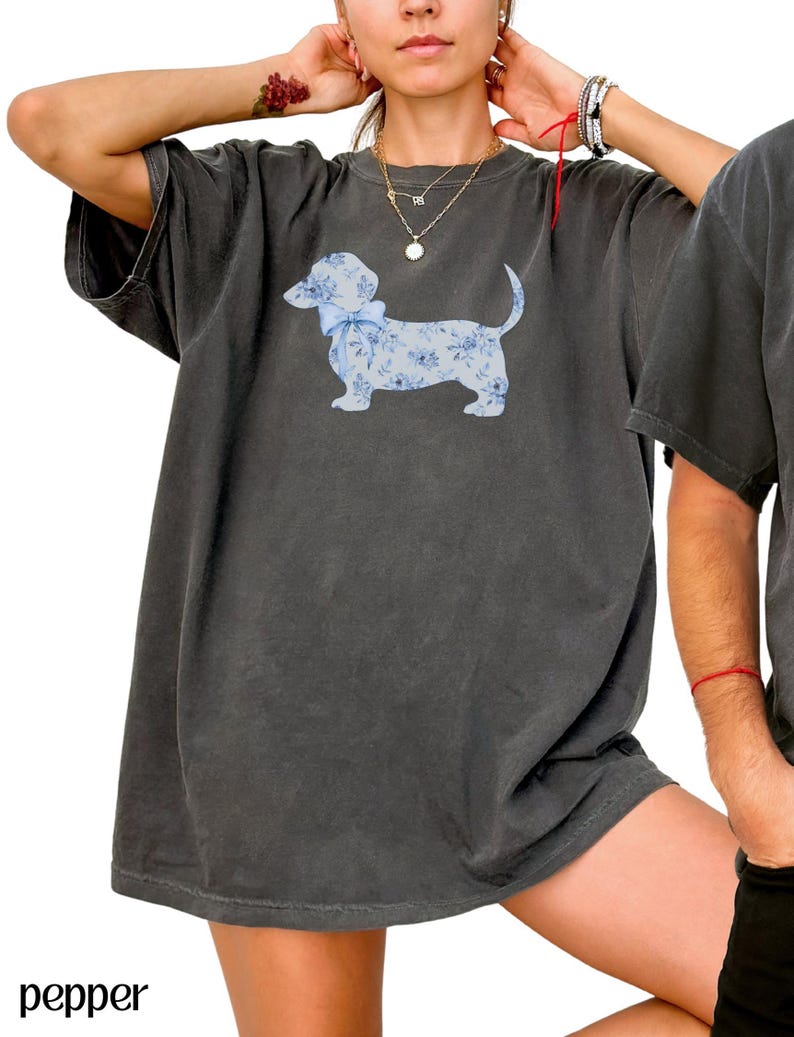May include: Dark gray t-shirt featuring a light blue dachshund illustration with a floral pattern and a bow. The word "pepper" is printed in the lower left corner of the image.