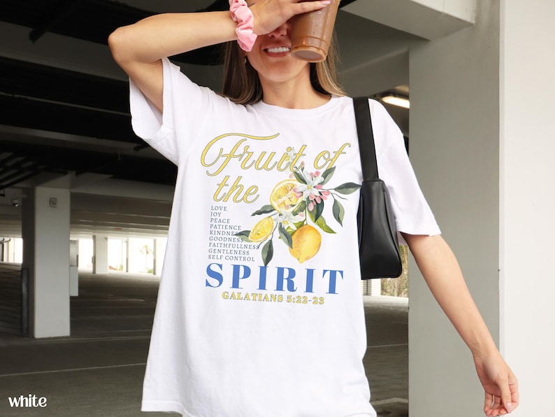 Op de afbeelding: Wit T-shirt met de tekst "Fruit of the Spirit" in geel en blauw, met een citroen- en bloemenontwerp. Het shirt vermeldt ook deugden als liefde, vreugde, vrede en geduld. De tekst "Galaten 5:22-23" is ook opgenomen.