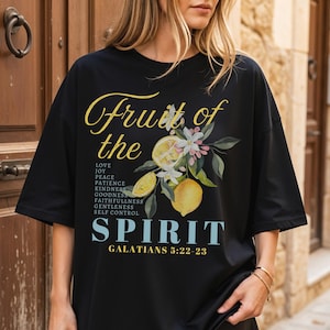 Op de afbeelding: Zwart T-shirt met een bloemen- en citroenontwerp. De tekst luidt "Vrucht van de Geest" met een lijst van deugden en de Bijbelverwijzing Galaten 5:22-23. Het woord "zwart" staat linksonder.