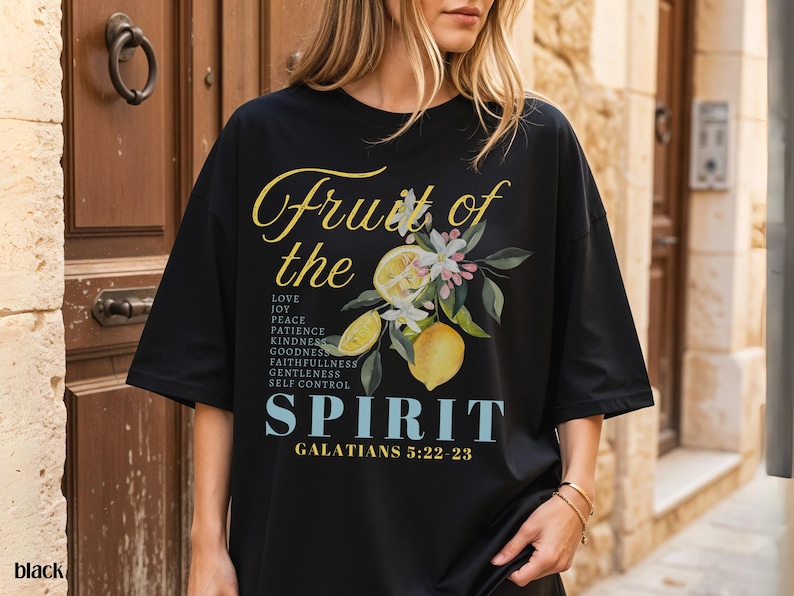Op de afbeelding: Zwart T-shirt met een bloemen- en citroenontwerp. De tekst luidt "Vrucht van de Geest" met een lijst van deugden en de Bijbelverwijzing Galaten 5:22-23. Het woord "zwart" staat linksonder.