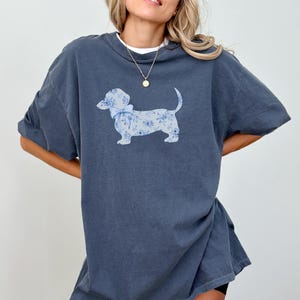 May include: Denim-colored t-shirt featuring a dachshund dog illustration. The dog is adorned with a blue floral pattern and a matching bow. The word "denim" is printed in the upper right corner. The shirt has short sleeves and a relaxed fit.