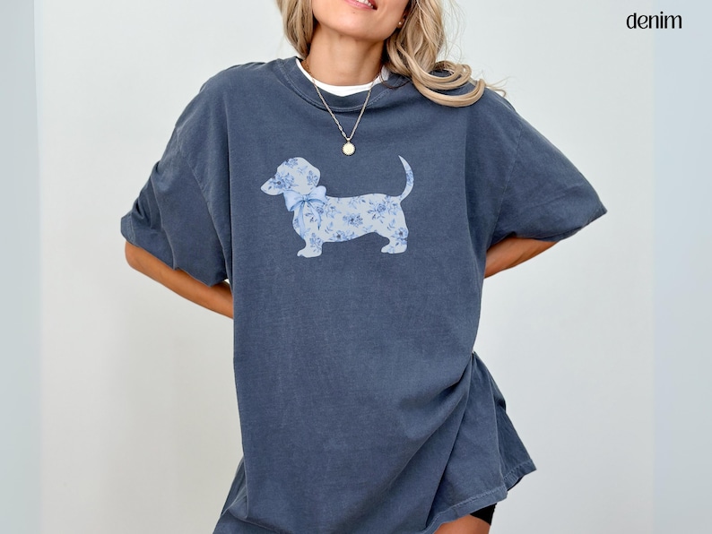May include: Denim-colored t-shirt featuring a dachshund dog illustration. The dog is adorned with a blue floral pattern and a matching bow. The word "denim" is printed in the upper right corner. The shirt has short sleeves and a relaxed fit.