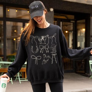 May include: Black sweatshirt with white line drawings of cowboy-themed elements, including bows, a horse and rider, a horseshoe, a cowboy hat, and a rope. The model is wearing a black cap with the number 11:11 on it.