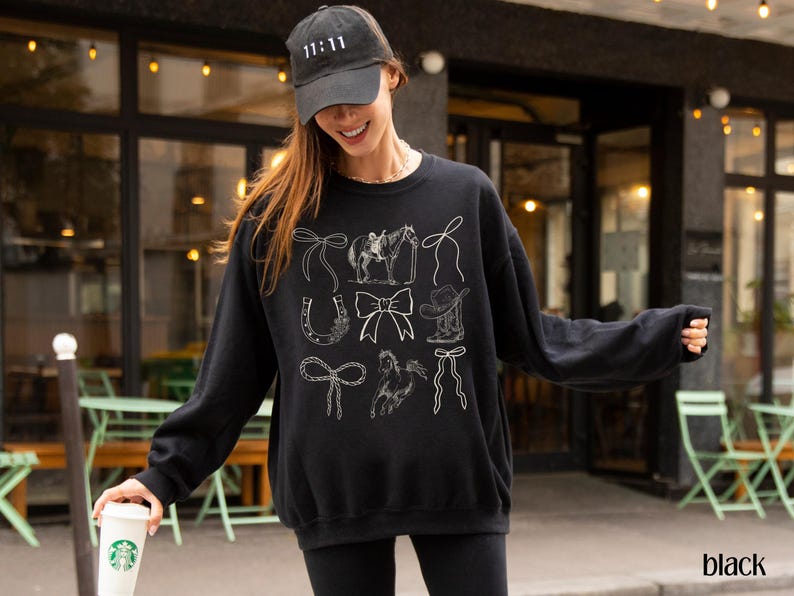 May include: Black sweatshirt with white line drawings of cowboy-themed elements, including bows, a horse and rider, a horseshoe, a cowboy hat, and a rope. The model is wearing a black cap with the number 11:11 on it.