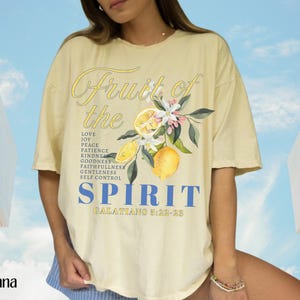 Op de afbeelding: Lichtgeel T-shirt met de woorden "Fruit of the Spirit" in script- en blokletters. Het shirt heeft een citroen- en bloemenafbeelding. De tekst bevat "Liefde, Vreugde, Vrede, Geduld, Vriendelijkheid, Goedheid, Trouw, Zachtmoedigheid, Zelfbeheersing, Galaten 5:22-23".