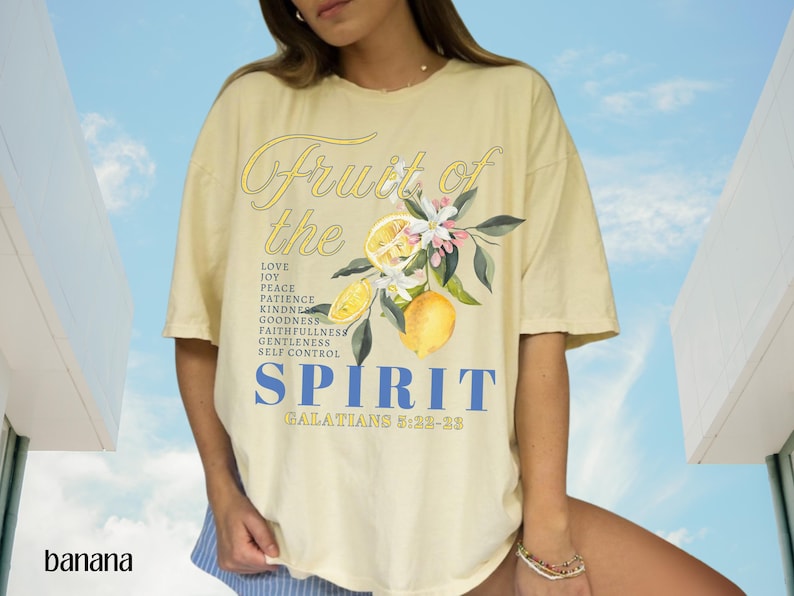 Op de afbeelding: Lichtgeel T-shirt met de woorden "Fruit of the Spirit" in script- en blokletters. Het shirt heeft een citroen- en bloemenafbeelding. De tekst bevat "Liefde, Vreugde, Vrede, Geduld, Vriendelijkheid, Goedheid, Trouw, Zachtmoedigheid, Zelfbeheersing, Galaten 5:22-23".