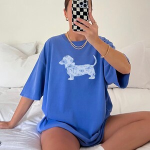 May include: A blue t-shirt with a floral dachshund graphic. The shirt is a solid blue color and has short sleeves. The text "flo blue" is at the bottom of the image.