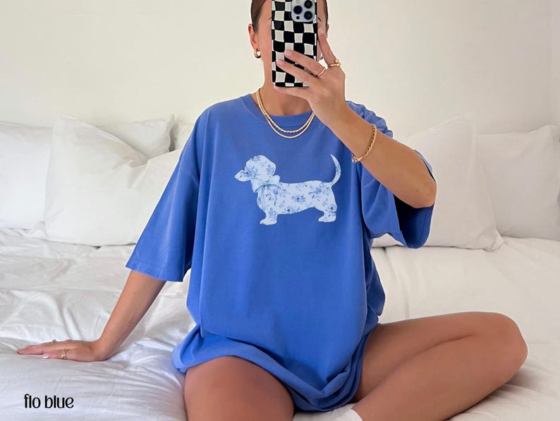 May include: A blue t-shirt with a floral dachshund graphic. The shirt is a solid blue color and has short sleeves. The text "flo blue" is at the bottom of the image.