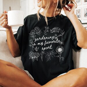 May include: Black t-shirt with the text "gardening is my favorite sport" in a white script font. The shirt features a floral design around the text. The person is holding a white mug.