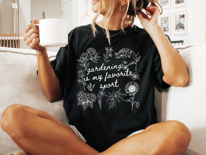 May include: Black t-shirt with the text "gardening is my favorite sport" in a white script font. The shirt features a floral design around the text. The person is holding a white mug.