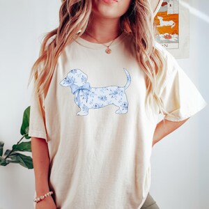 May include: Ivory-colored t-shirt featuring a dachshund dog design. The dog is adorned with a blue floral pattern and a matching bow. The shirt is a comfortable, casual style, perfect for everyday wear.