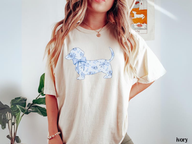 May include: Ivory-colored t-shirt featuring a dachshund dog design. The dog is adorned with a blue floral pattern and a matching bow. The shirt is a comfortable, casual style, perfect for everyday wear.