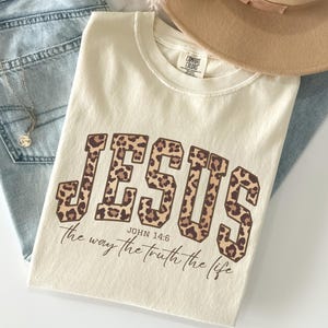 May include: Ivory-colored t-shirt with the word "JESUS" in a leopard print design. The text "John 14:6 the way the truth the life" is below the word "JESUS". The shirt is laid on top of blue jeans and a tan hat.