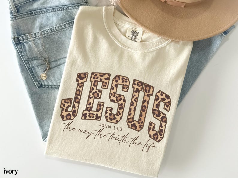 May include: Ivory-colored t-shirt with the word "JESUS" in a leopard print design. The text "John 14:6 the way the truth the life" is below the word "JESUS". The shirt is laid on top of blue jeans and a tan hat.