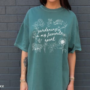 May include: Teal-colored t-shirt with the phrase "gardening is my favorite sport" in white script, surrounded by floral illustrations. The shirt is a relaxed fit and has short sleeves.