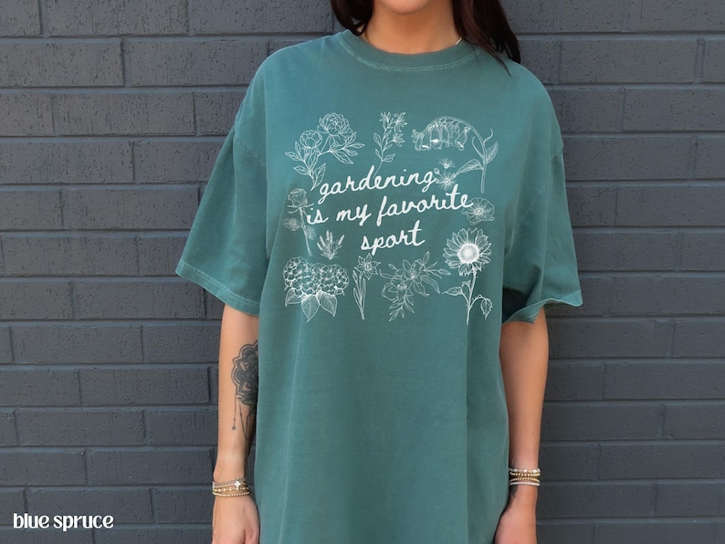 May include: Teal-colored t-shirt with the phrase "gardening is my favorite sport" in white script, surrounded by floral illustrations. The shirt is a relaxed fit and has short sleeves.