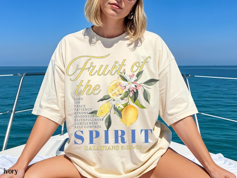 Op de afbeelding: Ivoor kleurig T-shirt met de tekst "Fruit of the Spirit" in gele letters en blauwe blokletters. Het shirt heeft een citroen en bloemen grafische afbeelding. De tekst bevat een lijst met deugden en de Bijbeltekst Galaten 5:22-23.