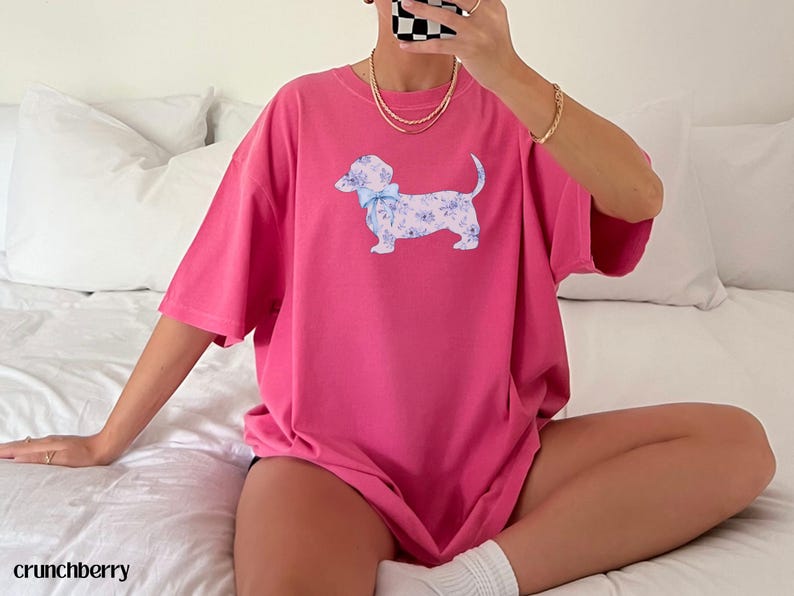 May include: A hot pink t-shirt featuring a dachshund dog illustration with a floral pattern and a blue bow. The dog is white with a blue floral design. The shirt is oversized and has short sleeves.