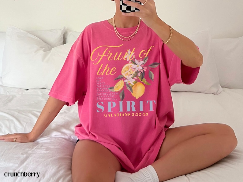 Op de afbeelding: Een felroze t-shirt met de tekst "Fruit of the Spirit" en een bloemen- en citroenafbeelding. Het shirt bevat ook de Bijbelverwijzing Galaten 5:22-23. Het shirt heeft een relaxte pasvorm.